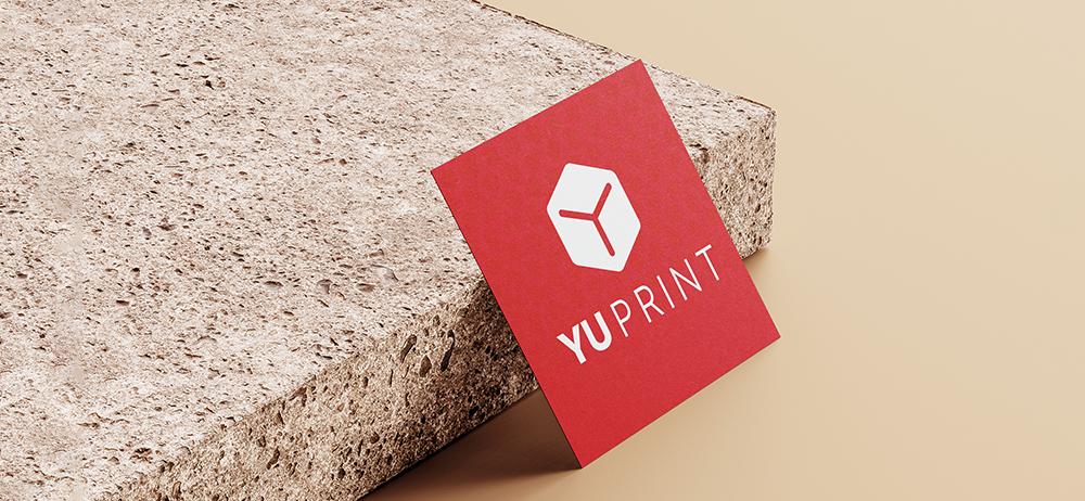 Tarjeta cuadrada roja con logotipo de Yuprint apoyada sobre superficie de piedra.
