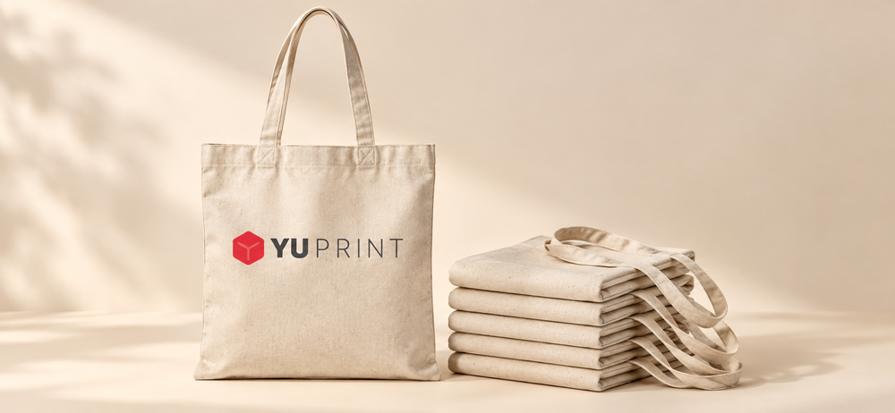 Bolsa de algodón personalizada con logo de Yuprint junto a un lote de tote bags plegadas